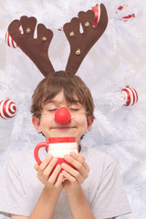 Kid wearing reindeer antlers and red nose smelling a mug of hot chocolate