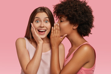 Young black woman whispers secret to her Caucasian friend, gossip together and spread rumours. Emotional European girl feels amazed to hear confidential information from companion. Secrecy, gossiping - obrazy, fototapety, plakaty