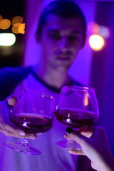 blurred young handsome man with close up  glass of red wine and female hand with wine  in the evening in the light of neon lamps