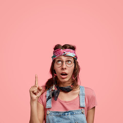 Only look up. Surprised fashionable hipster impressed by something awesome, points with index finger upwards, dressed in stylish dungarees, shows free space over pink background. Subculture.