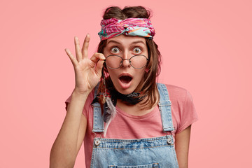 Emotional surprised young hippy woman keeps hand on rim of round glasses, wears fashionable...