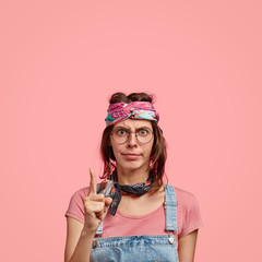 Photo of serious angy hippie being discontent with noisy neighbours, points with index finger upwards, wears stylish headband and denim overalls, free space against pink background for your text