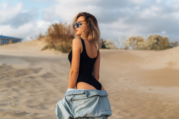 Sexy blonde girl in a black swimsuit and blue sunglasses posing on the beach. Beautiful model with perfect figure sunbathes and rests. Travel concept.