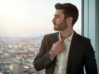 Portrait of stylish young man wearing business suit, standing next to window overlooking modern...