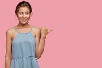 Studio shot of pleased European woman with joyful expression, enjoys watching cool performance, dressed in fashionable dress, isolated over pink background. Advertisement and promotion concept