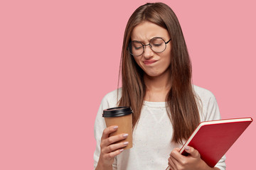 Photo of pretty dark haired woman frowns face in displeasure holds takeaway coffee, doesnt want drink anymore, carries textbook, wears casual outfit, isolated over pink background with copy space