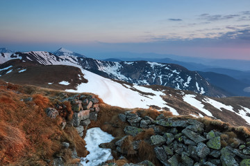 The landscape of the high mountains in snow. The grass and the rocks on the meadow. Sunrise is lightening the horizon. Sky with clouds.