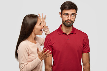 Young family couple have conflict. Angry brunette young European woman gestures with hands, shouts at husband who is guilty, stands together against white background, have dispute and quarrel