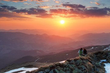 There is a couple of friends sitting on the lawn full of rocks. The landscape of the mountains in fog. Sunset. The sky with clouds in beautiful light.