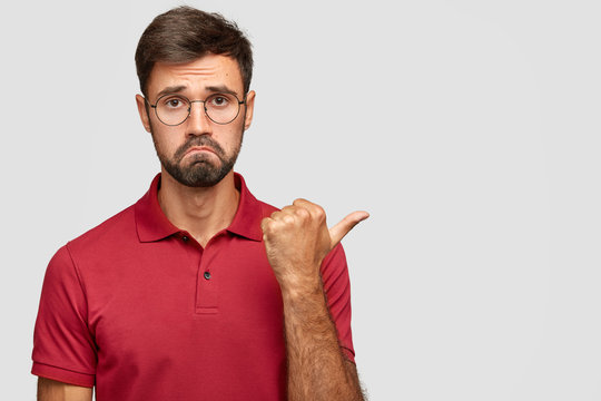 Image of unhappy Caucasian man feels regret and envy, sulks face, has gloomy expression, points with thumb aside, dressed in red casual t shirt, isolated over white wall, free space for your promotion