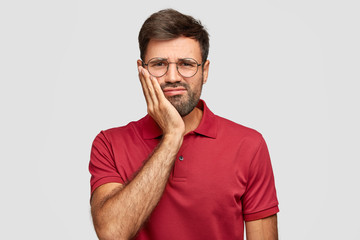 Handsome man with dark stubble, grimaces from painful feeling, has decay, suffers from terrible toothache, waits for dentist, dressed in casual red t shirt poses over white background. Dental problems