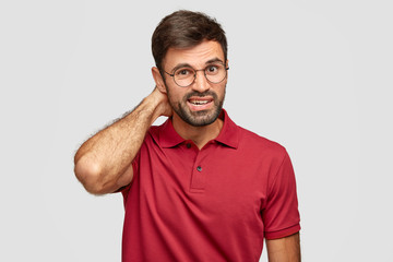 Photo of puzzled man with stubble, scratches head, looks in bewilderment, wears round spectacles for good vision, dressed in casual red t shirt, models against white background has confused expression