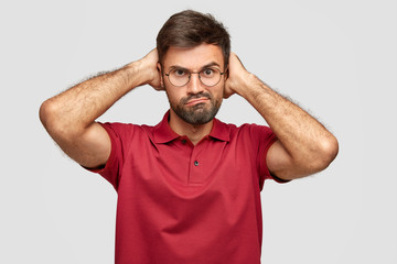 Photo of displeased unshaven man frowns face and looks angrily, purses lips, keeps hands behind...
