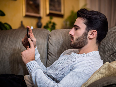 Handsome young man at home, reading with ebook reader lying on a couch