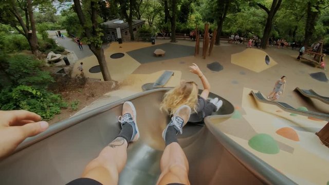 Young couple or teenage friends, pov shot going down metal kids or playground slide at park during summer vacation, fun and unconventional activity for weekend or date, laugh and smile