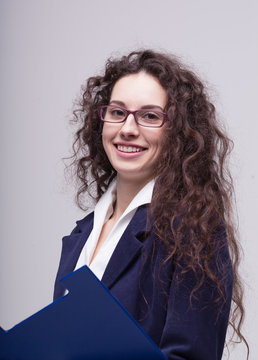 Young businesswoman holding a clipboard. Portrait of cheerful smiling businesswoman in glasses, with blue folder, posing at studio, over gray background