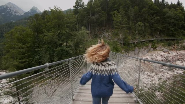Beautiful slow motion shot of back of young blonde woman in authentic native blue wool sweater run into forest on hanging wooden bridge, concept freedom and adventure travel lifestyle in nature