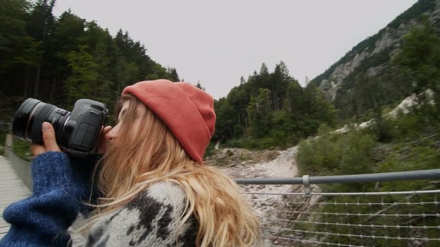 Camera goes around young hipster or millennial woman in authentic blue woolen sweater, trendy outfit walks on wooden bridge in high alps, creates content for social media or nature magazine