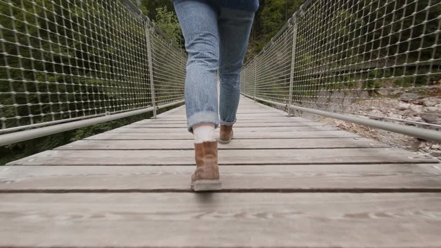 Close up shot of woman in blue denim jeans and brown leather heavy duty hiking boots walks on wooden bridge hanging over river in mountain forest, adventure seeker urban nomad