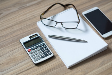 Notepad, calculator, smartphone, glasses and sliver ballpen on wooden office desk. Selective focus.