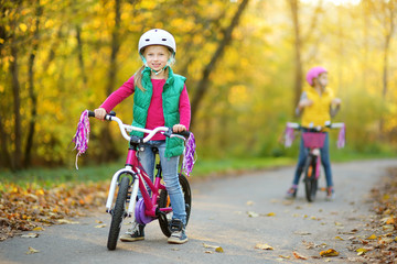 Naklejka premium Cute sisters riding bikes in a city park on sunny autumn day. Active family leisure with kids. Children wearing safety hemet while riding a bicycle.