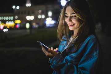 Blogger hipster using in hands gadget mobile phone. Woman pointing finger on blank screen smartphone on background bokeh light in night atmospheric city street.