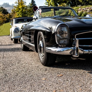 Schwarzer Oldtimer Cabrio mit offenen Dach dahinter wei&szlig;e Oldtimerlimusine mit geschlossenen Verdeck parken an einem Schotterweg
