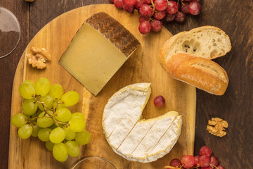 An overhead photo of cheeses, Camembert and a cured cheese, with grapes and nuts on a dark rustic wooden background