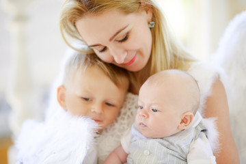 Beautiful mother with her toddler and newborn sons wearing angel costumes. Cheerful moment, loving family.