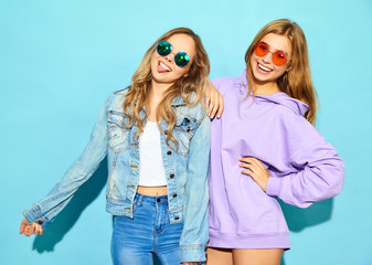 Two young beautiful blond smiling hipster girls in trendy summer clothes. Sexy carefree women posing near blue wall in sunglasses. Positive models going crazy