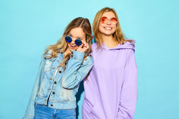 Two young beautiful blond smiling hipster girls in trendy summer clothes. Sexy carefree women posing near blue wall in sunglasses. Positive models going crazy and hugging