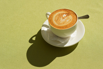 A Cup of cappuccino with a pattern on the table lit by the morning sun.