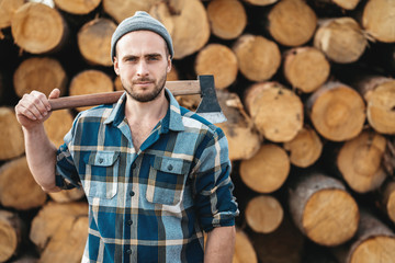 Strong bearded lumberman wearing plaid shirt holds axe on his shoulder on background of sawmill
