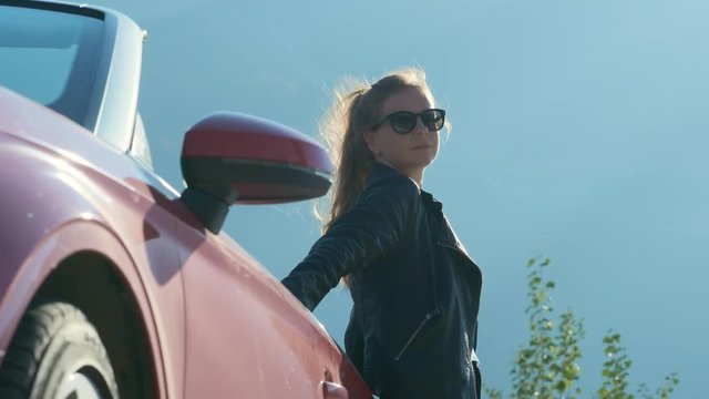 sexy young woman in black leather jacket posing with red cabriolet against sky background.