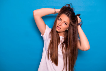Obraz na płótnie Canvas Cute brunette woman with long hair posing in white t-shirt on a blue background. Emotional portrait. She tries to tear her hair