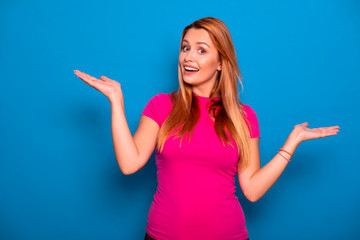 Fototapeta premium Sexy plus size model with long red hair in pink t-shirt on a blue background. Emotional portrait. She try to decide what to choose, variants on her hands