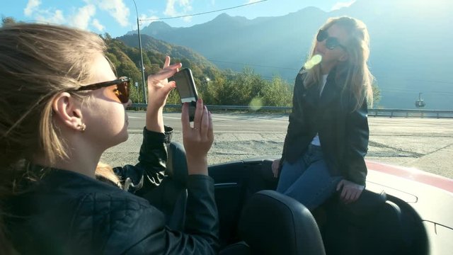Two young beautiful girls in black glasses and leather jackets in a red cabriolet near the high mountains. They make a photo session, take pictures of themselves on a smartphone, have fun