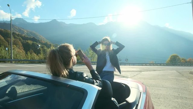 Two young beautiful girls in black glasses and leather jackets in a red cabriolet near the high mountains. They make a photo session, take pictures of themselves on a smartphone, have fun