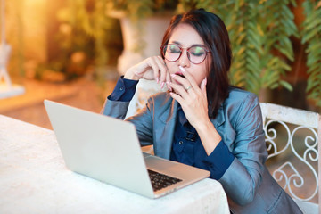 cute and charming businesswoman with glasses working on laptop computer and yawning in restaurant or cafe