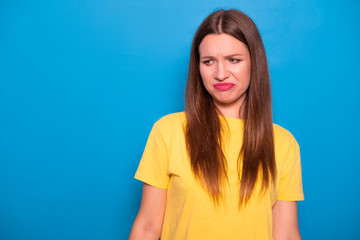 Cute brunette woman with long hair posing in yellow t-shirt on a blue background. Emotional portrait. She shows her disgust