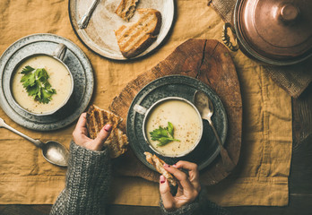 Autumn, Winter home dinner. Flat-lay of Fall warming celery cream soup and female hands with grilled bread over linen tablecloth, top view. Comfort food, vegetarian, healthy and slow food concept