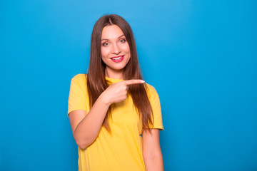 Cute brunette woman with long hair posing in yellow t-shirt on a blue background. Emotional portrait. She smiles happily with flawless white teeth and poins finger on something