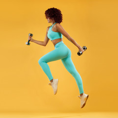 Fototapeta na wymiar Strong athletic, woman sprinter or runner, running on yellow background with dumbbells wearing sportswear. Fitness and sport motivation.