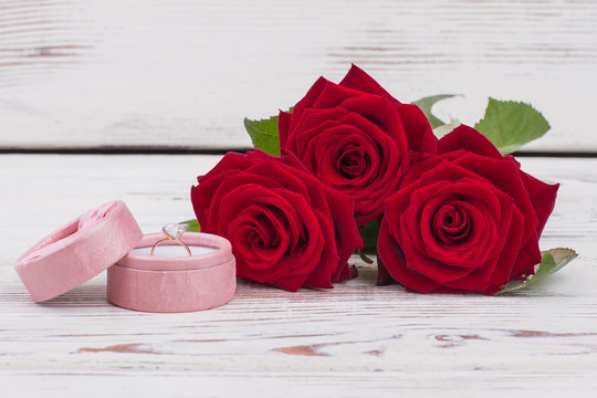 Red roses and gift box with golden ring. Valentines Day background with a bunch of blooming roses and engagement ring on white wooden background.