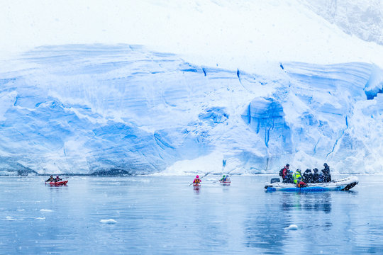 Snowfall over the motor boat with tourists and kayaks in the bay with huge blue glacier wall in the background, near Almirante Brown, Antarctic peninsula