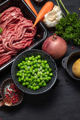 Raw minced meat in container surrounded by ingredients for shepherds pie , green peas, carrot, onion and seasonings, on black background, top view. Vertical composition