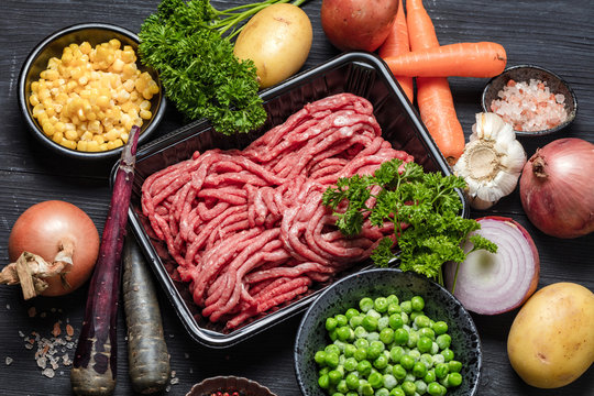 Raw minced meat in container surrounded by ingredients for shepherds pie , green peas, yellow corn, carrot, onion and seasonings, on black background, top view. Horizontal composition