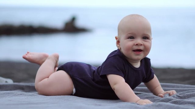 The baby lies on his stomach on the black sand near the ocean and laughs looking at the camera