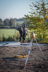 Construction workers measuring height at a site, with earth level equipment in the foreground. 