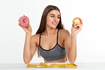 Diet concept. Beautiful slim girl with a smile, chooses between a donut and a fresh apple. Studio, white background.
